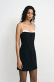A model wearing a black mini dress with a white contrast panel at the neckline, showing the pleated front detail, turned slightly to the side.