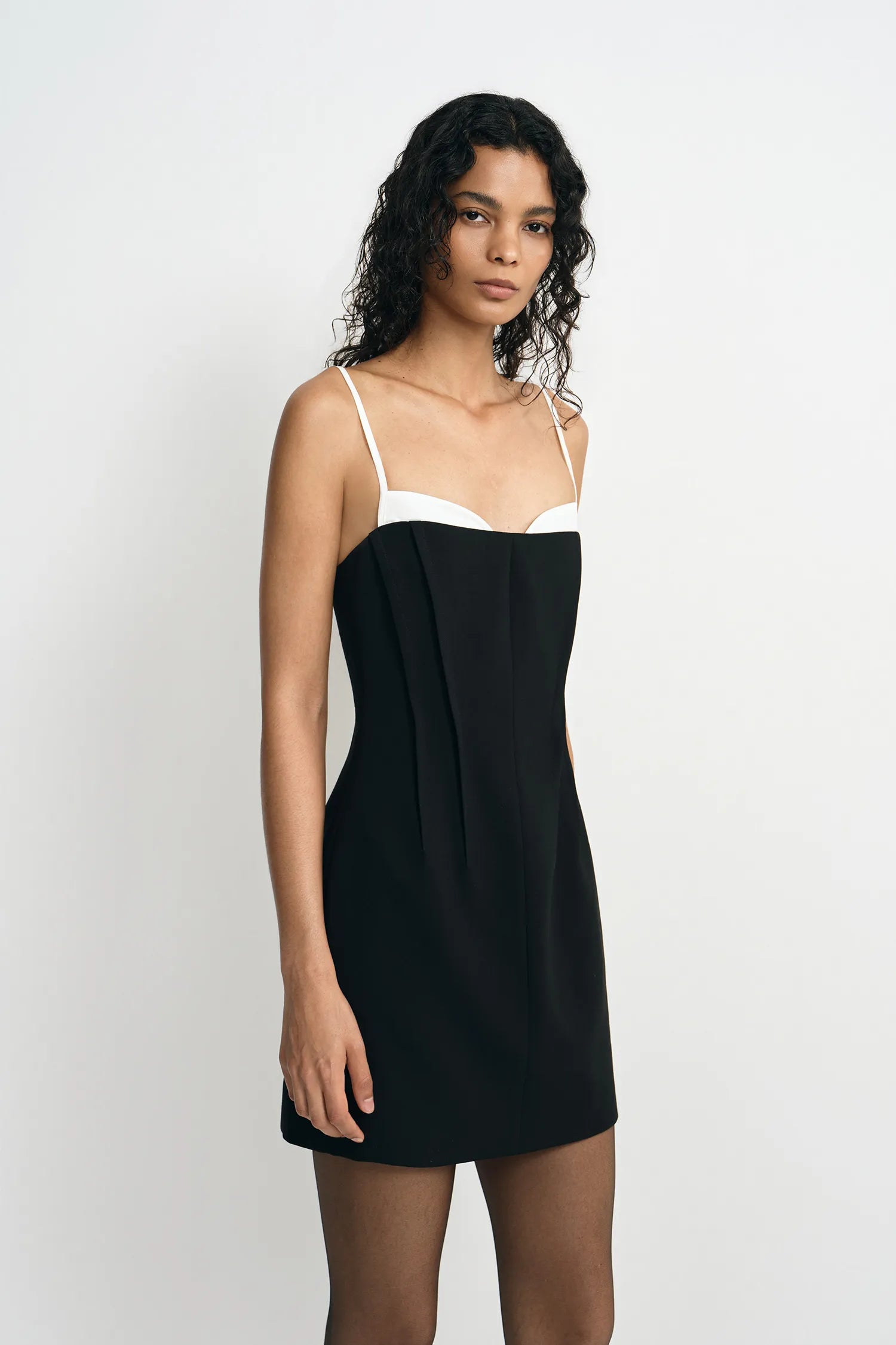 A model wearing a black mini dress with a white contrast panel at the neckline, showing the pleated front detail, turned slightly to the side.