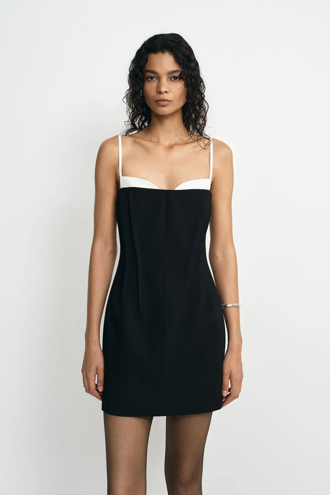 A model wearing a black mini dress with a white contrast panel at the neckline and thin straps, facing the camera.