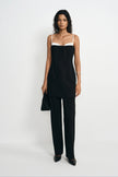 A model stands against a plain white studio background wearing a black camisole-style top with a white band at the neckline and matching black wide-leg pants, holding a black clutch.
