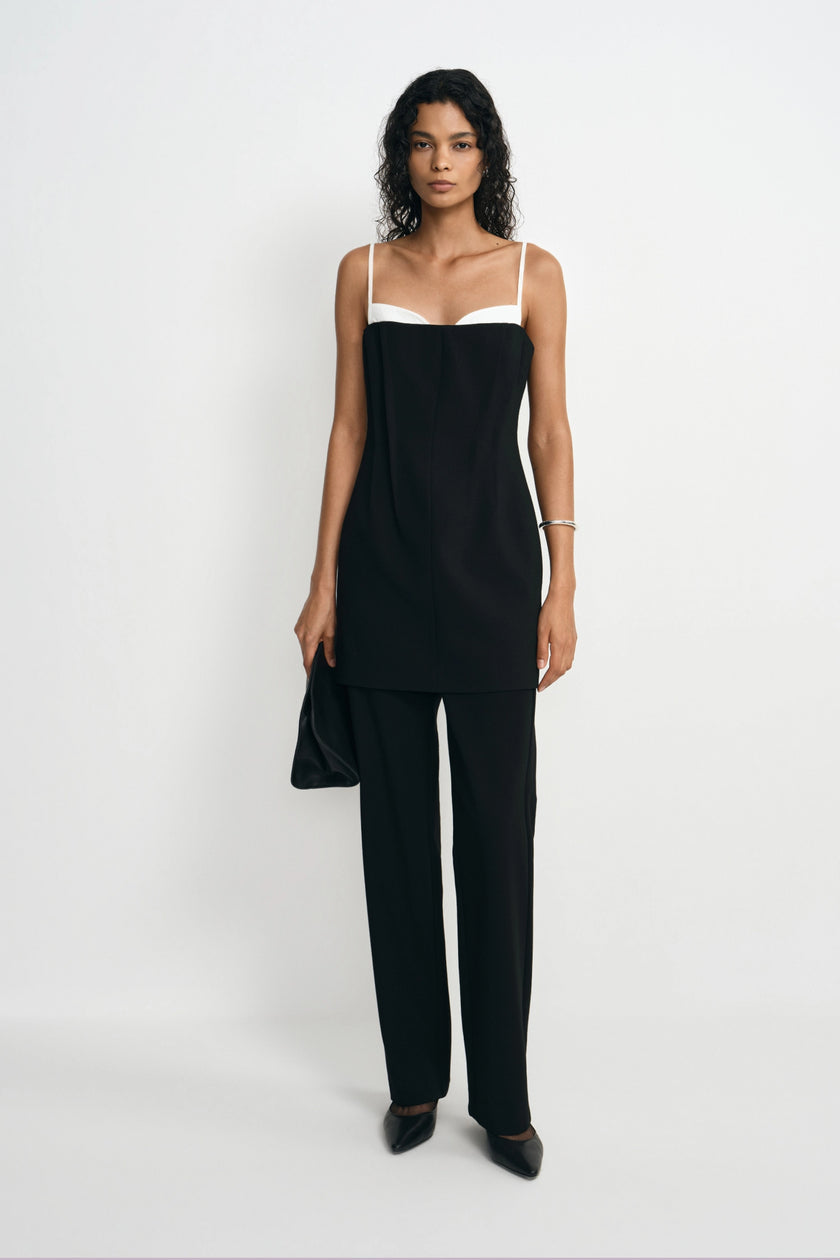 A model stands against a plain white studio background wearing a black camisole-style top with a white band at the neckline and matching black wide-leg pants, holding a black clutch.