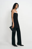 A model wearing a black sleeveless jumpsuit with a white underband and wide legs, holding a black clutch bag against a plain white studio background.