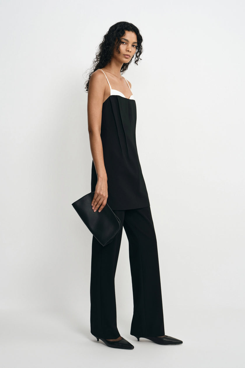 A model wearing a black sleeveless jumpsuit with a white underband and wide legs, holding a black clutch bag against a plain white studio background.