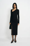 A model wearing a black long-sleeve asymmetrical wrap dress with a ruched skirt and thigh-high slit, paired with black pointed-toe flats and holding a black clutch, set against a plain white studio background.