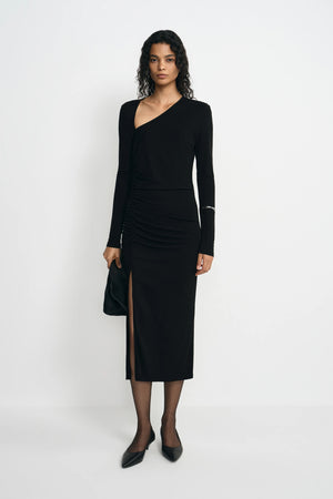 A model wearing a black long-sleeve asymmetrical wrap dress with a ruched skirt and thigh-high slit, paired with black pointed-toe flats and holding a black clutch, set against a plain white studio background.