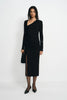 A model wearing a black long-sleeve asymmetrical wrap dress with a ruched skirt and thigh-high slit, paired with black pointed-toe flats and holding a black clutch, set against a plain white studio background.