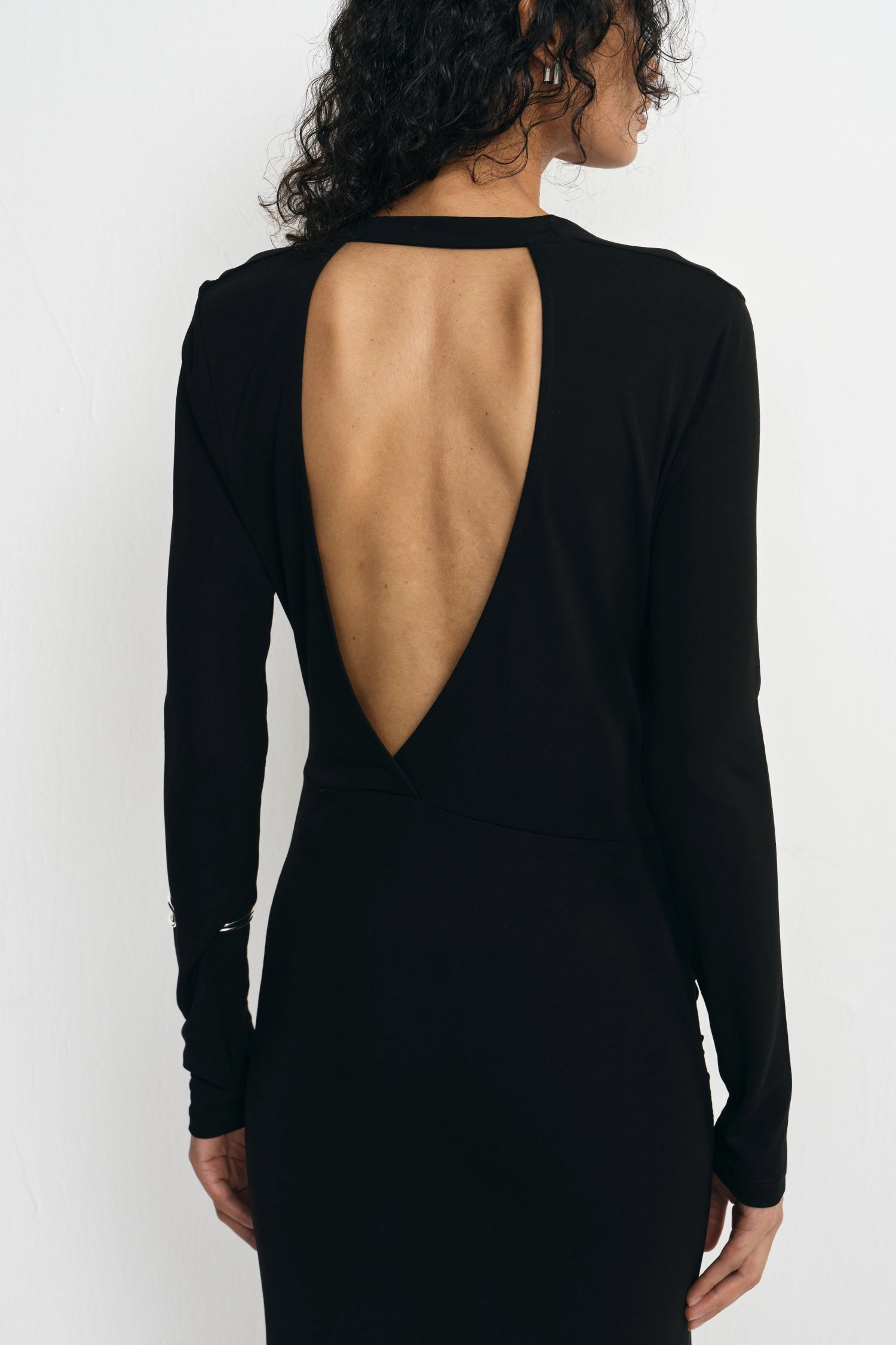 A woman shown from the back wearing a black long-sleeve dress with a large open back cutout, photographed against a plain light background.