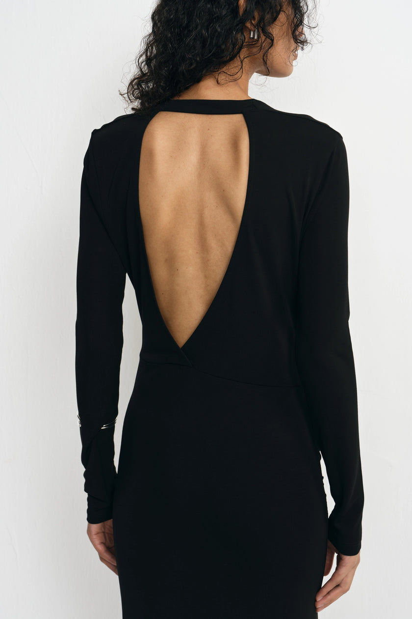 A woman shown from the back wearing a black long-sleeve dress with a large open back cutout, photographed against a plain light background.