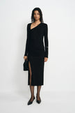 A model stands against a plain white studio background wearing a black long-sleeve asymmetrical wrap dress with a thigh-high slit, paired with black pointed shoes and holding a black clutch.