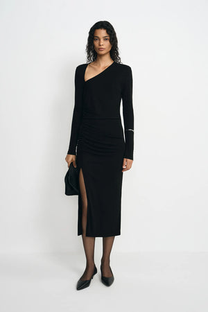 A model stands against a plain white studio background wearing a black long-sleeve asymmetrical wrap dress with a thigh-high slit, paired with black pointed shoes and holding a black clutch.