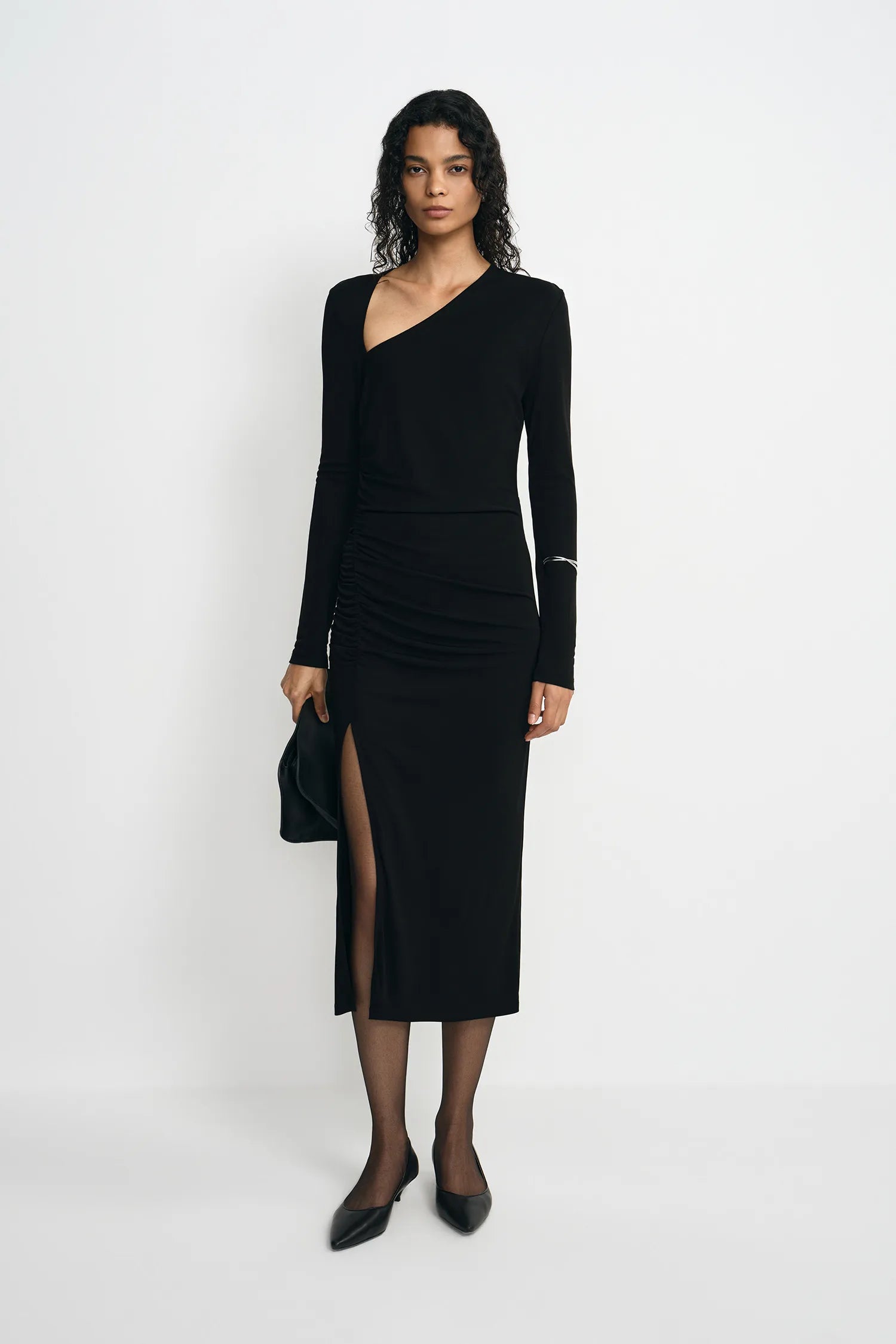 A model stands against a plain white studio background wearing a black long-sleeve asymmetrical wrap dress with a thigh-high slit, paired with black pointed shoes and holding a black clutch.