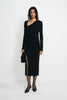A model stands against a plain white studio background wearing a black long-sleeve asymmetrical wrap dress with a thigh-high slit, paired with black pointed shoes and holding a black clutch.
