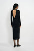 A woman viewed from the back wearing a black long-sleeve midi dress with a large open back, paired with sheer black tights and black pointed heels, against a plain light gray studio background.