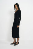 A woman modeling a long-sleeve black wrap-style midi dress with a V-neckline and side slit, paired with black pointed flats, against a plain light gray studio background.