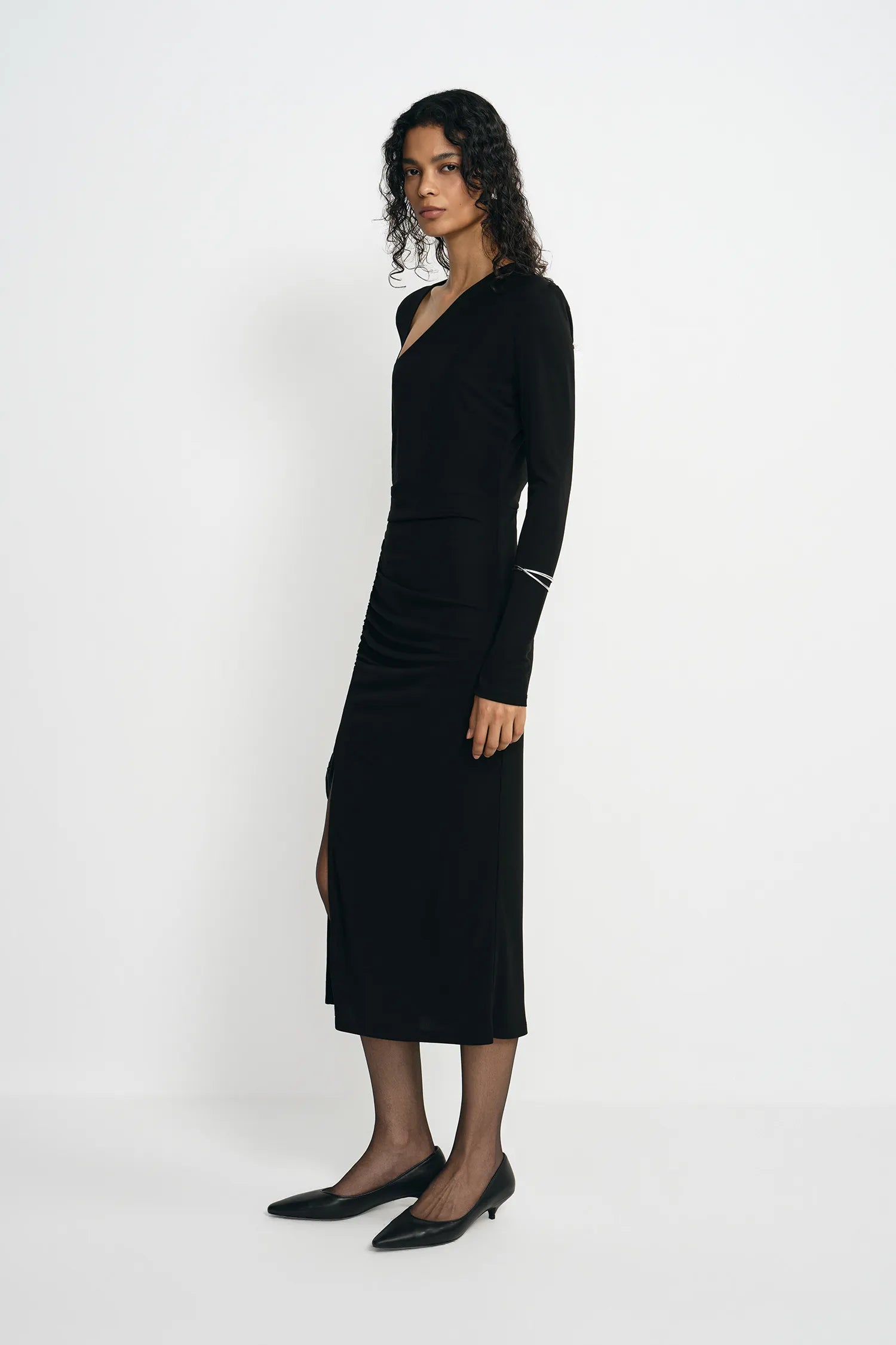 A woman modeling a long-sleeve black wrap-style midi dress with a V-neckline and side slit, paired with black pointed flats, against a plain light gray studio background.