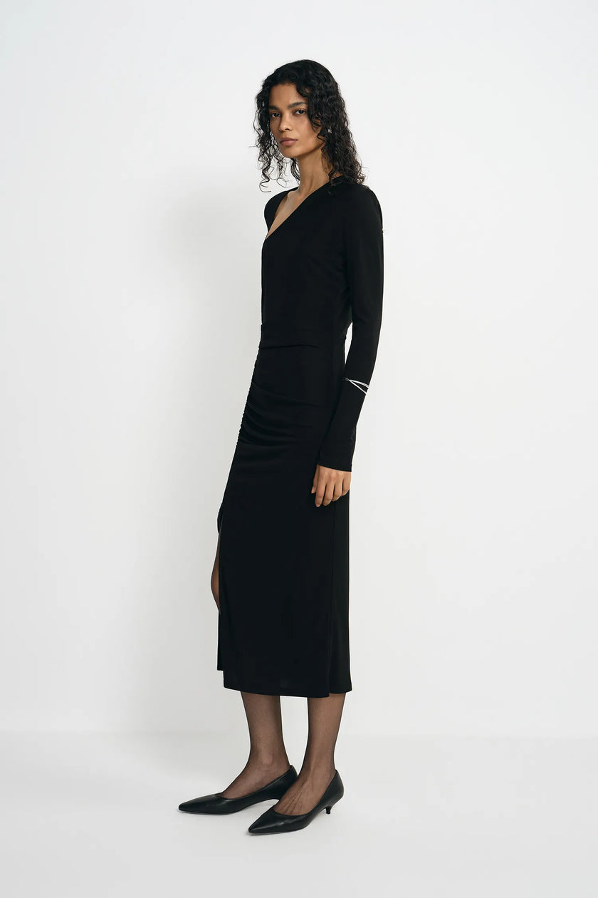 A woman modeling a long-sleeve black wrap-style midi dress with a V-neckline and side slit, paired with black pointed flats, against a plain light gray studio background.