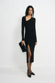 A model stands against a plain white studio background wearing a fitted black long-sleeve wrap-style midi dress with an asymmetrical neckline and a thigh-high front slit. She is also wearing black pointed shoes and holding a black clutch.