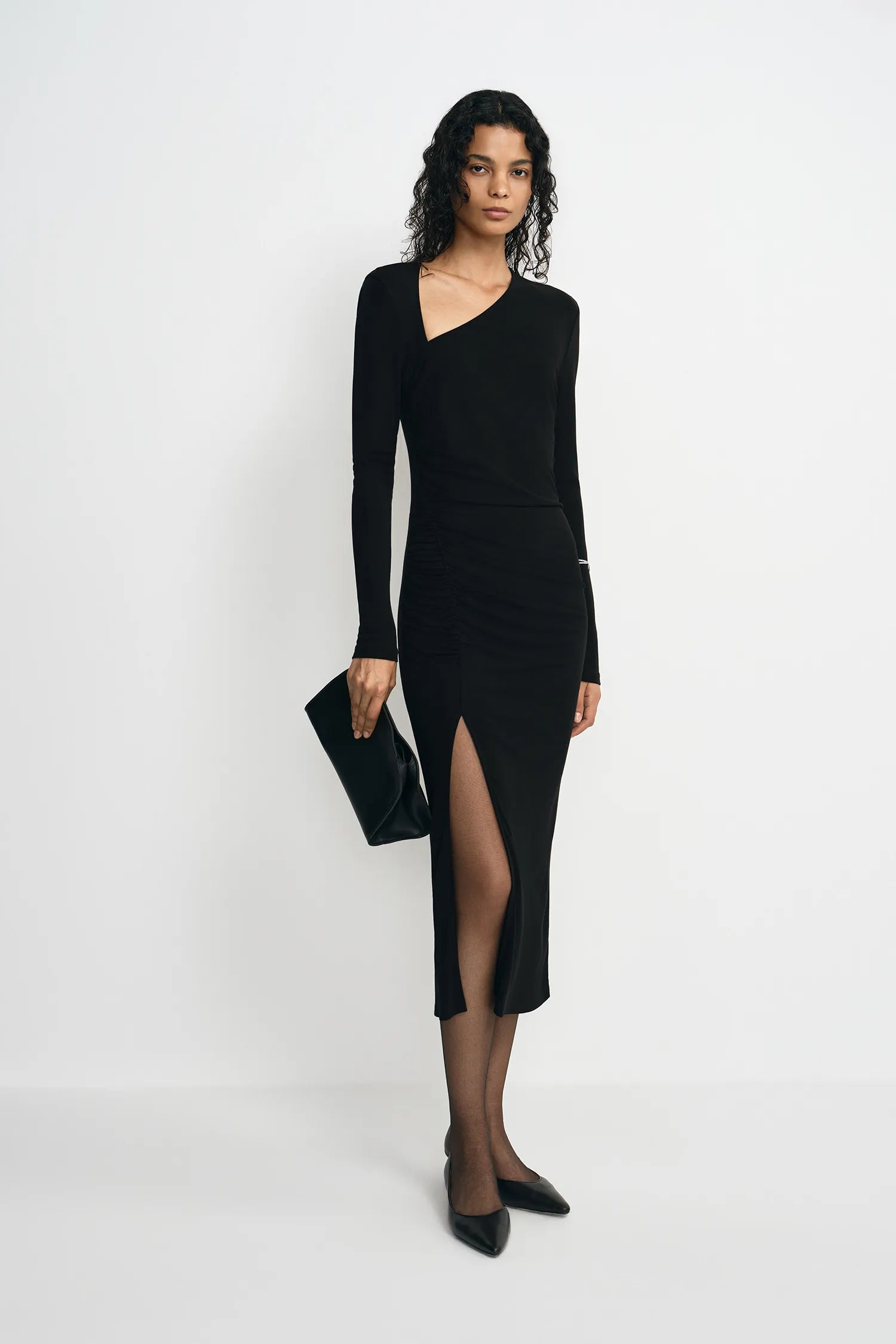 A model stands against a plain white studio background wearing a fitted black long-sleeve wrap-style midi dress with an asymmetrical neckline and a thigh-high front slit. She is also wearing black pointed shoes and holding a black clutch.