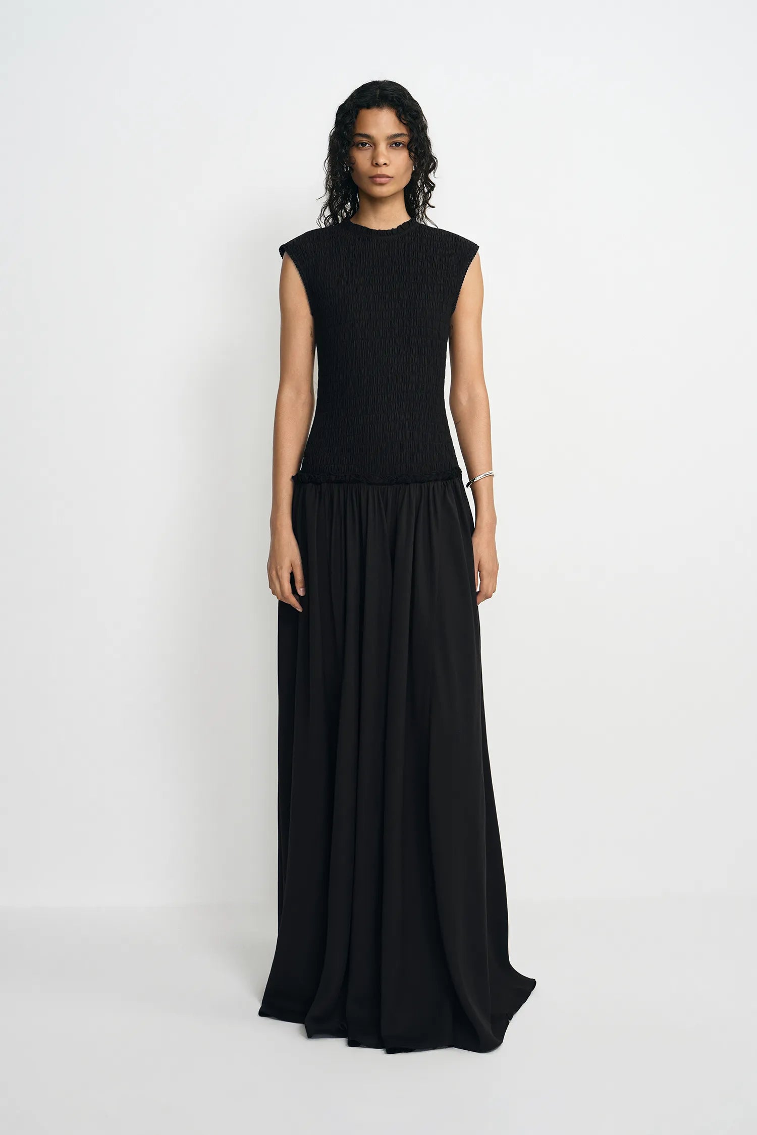 A model wearing a sleeveless black maxi dress with a textured knit bodice and a flowing chiffon skirt, standing against a plain light gray studio background.