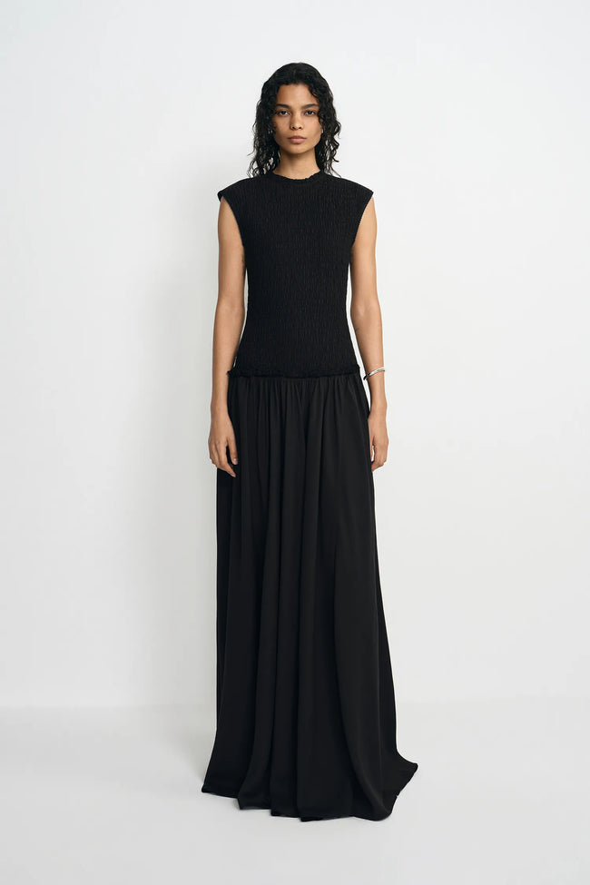 A model wearing a sleeveless black maxi dress with a textured knit bodice and a flowing chiffon skirt, standing against a plain light gray studio background.