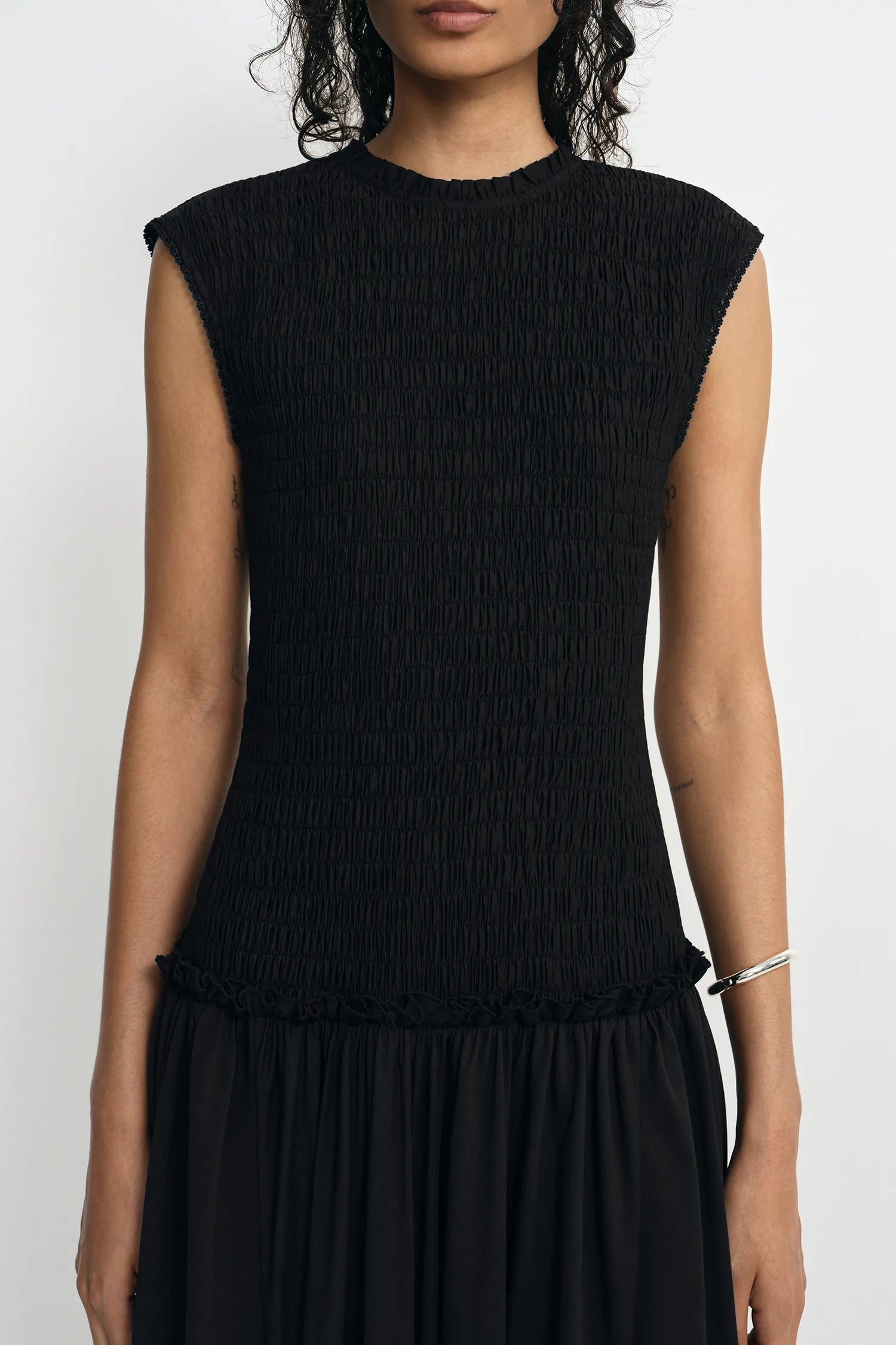A woman wearing a black sleeveless top with a smocked or crinkled texture and a ruffled hem, paired with a black skirt, against a plain light background.