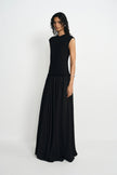 A woman modeling a sleeveless black maxi dress with a textured bodice and a flowing pleated skirt, standing against a plain light gray studio background.