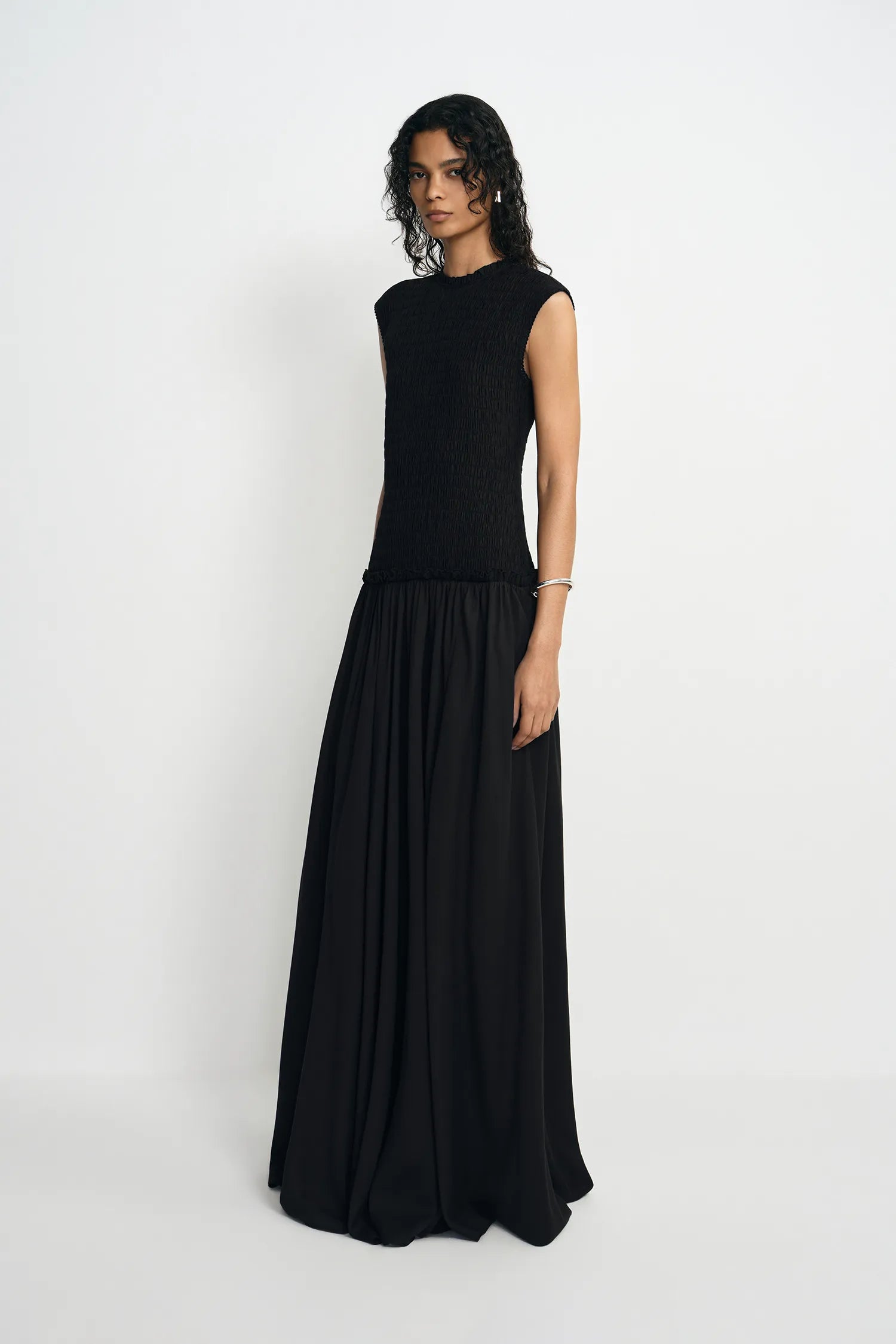 A woman modeling a sleeveless black maxi dress with a textured bodice and a flowing pleated skirt, standing against a plain light gray studio background.
