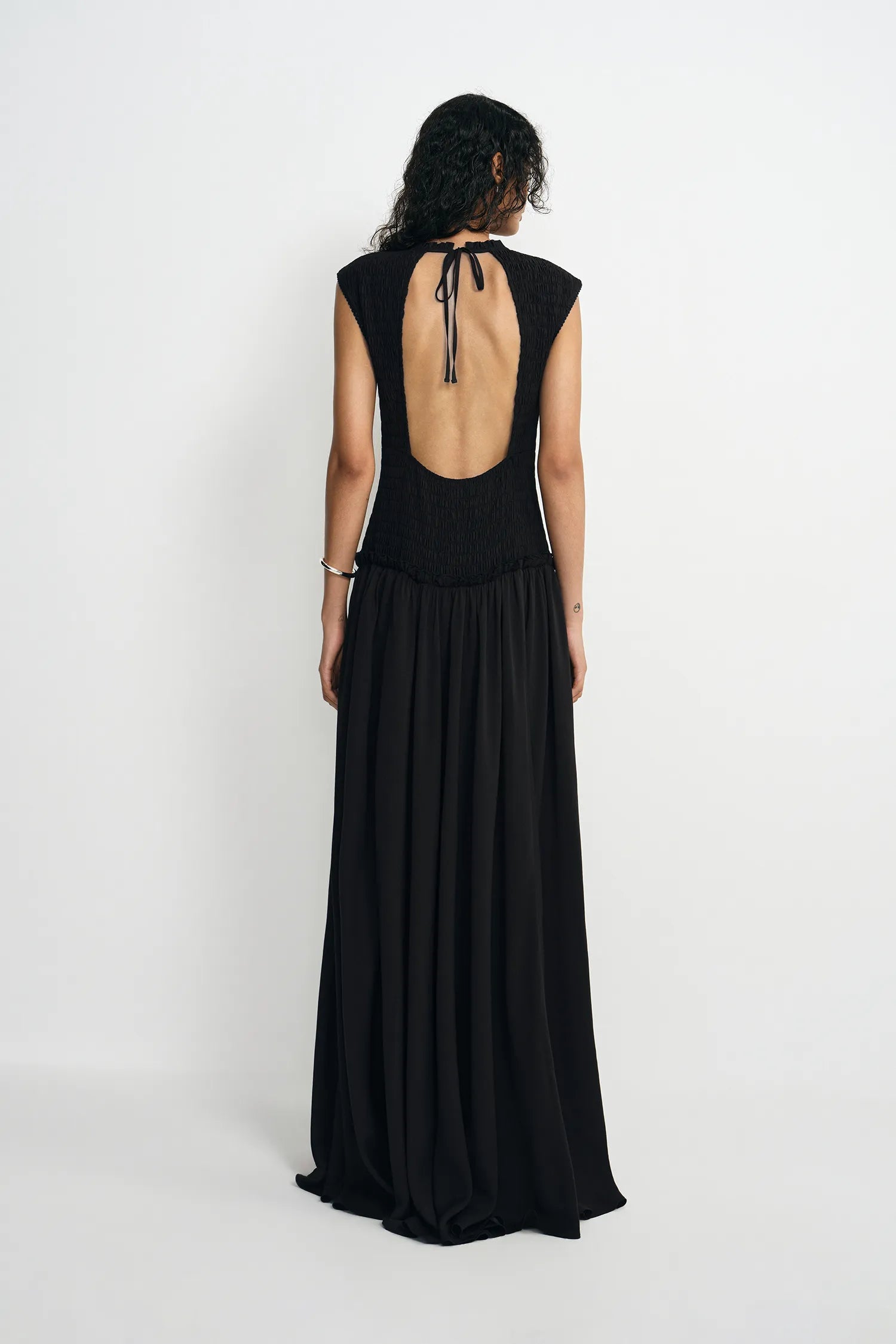 A woman viewed from the back wearing a sleeveless black maxi dress with a deep open back and tie detail, standing against a plain light gray studio background.