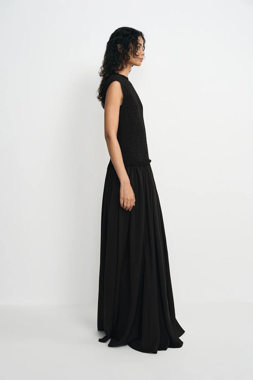 A woman modeling a sleeveless black maxi dress with a textured bodice and flowing skirt, shown in profile against a plain white studio background.