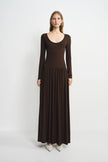 A woman modeling a long-sleeve, scoop-neck maxi dress in dark brown, paired with light gray pointed-toe heels, against a plain light gray studio background.