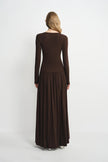 A woman viewed from the back wearing a long-sleeve, dark brown maxi dress with a fitted bodice and a flowing pleated skirt, set against a plain light gray studio background.