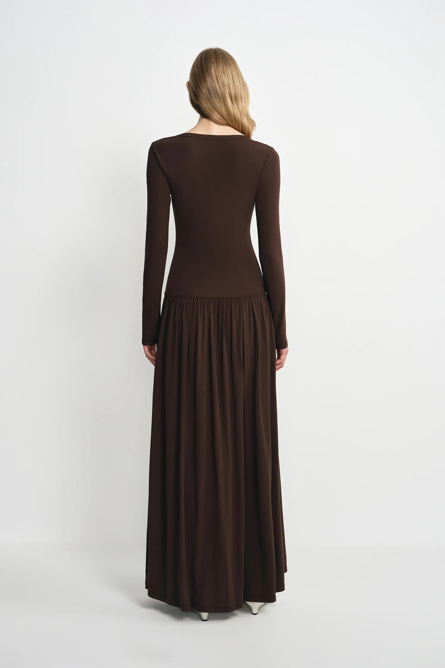 A woman viewed from the back wearing a long-sleeve, dark brown maxi dress with a fitted bodice and a flowing pleated skirt, set against a plain light gray studio background.