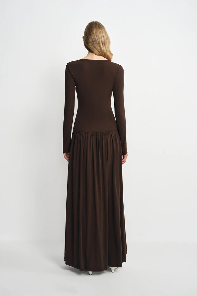 A woman viewed from the back wearing a long-sleeve, dark brown maxi dress with a fitted bodice and a flowing pleated skirt, set against a plain light gray studio background.