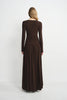 A woman viewed from the back wearing a long-sleeve, dark brown maxi dress with a fitted bodice and a flowing pleated skirt, set against a plain light gray studio background.