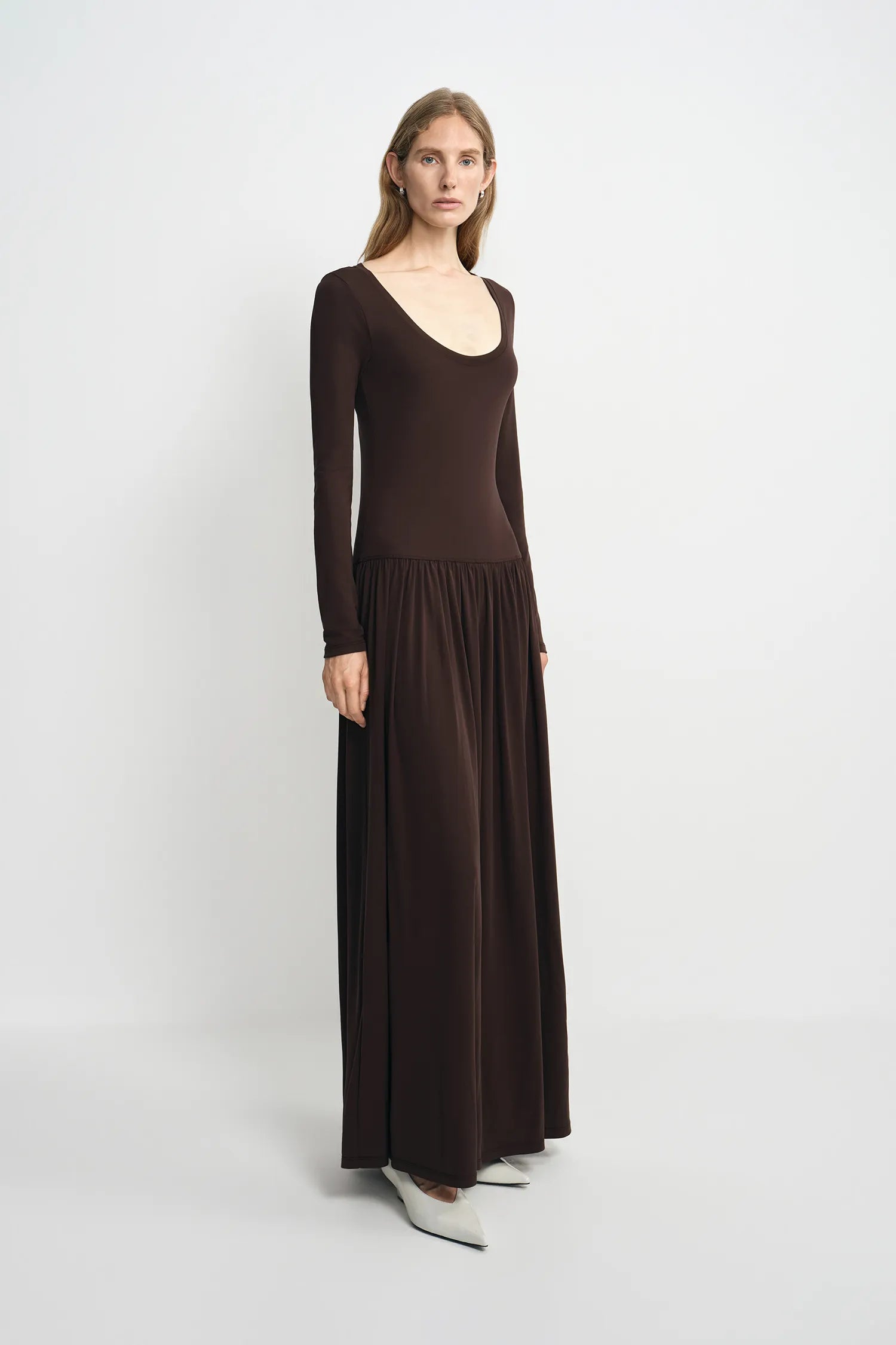 A woman modeling a dark brown long-sleeve maxi dress with a scoop neckline and a flowing skirt, standing against a light gray studio background.