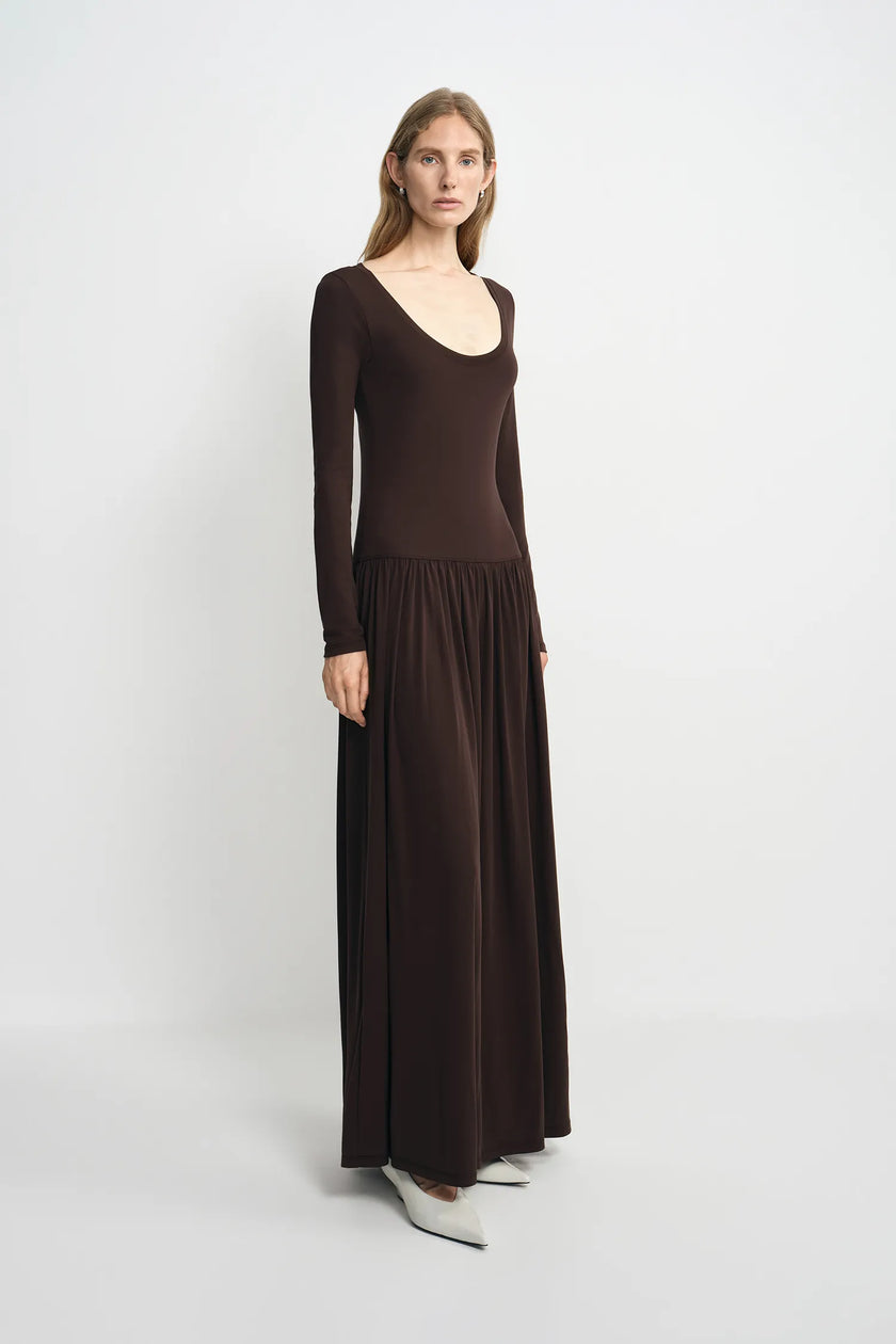 A woman modeling a dark brown long-sleeve maxi dress with a scoop neckline and a flowing skirt, standing against a light gray studio background.
