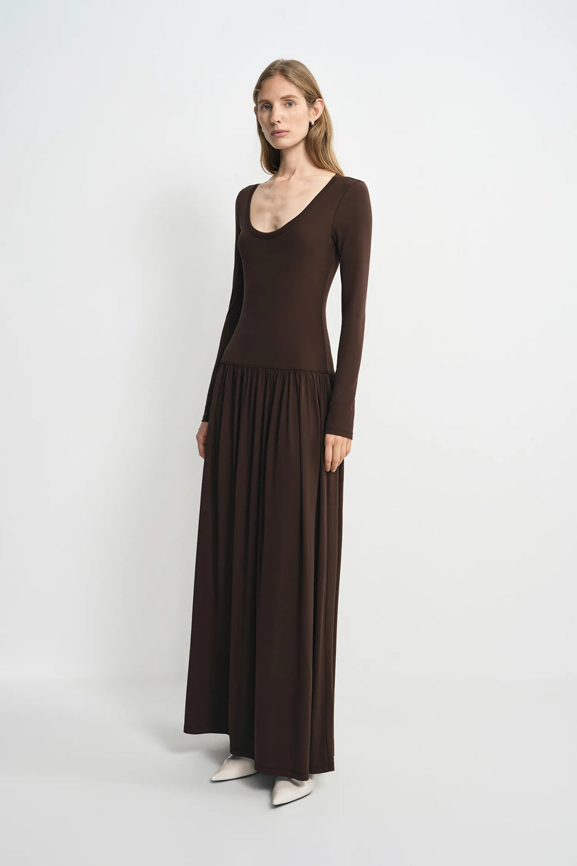 A woman modeling a long-sleeve, scoop-neck maxi dress in dark brown, paired with white pointed-toe flats, against a plain light gray studio background.