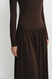 A close-up of a model wearing a dark brown long-sleeve maxi dress with a fitted bodice and a gathered, flowing skirt, shown against a plain light gray background.