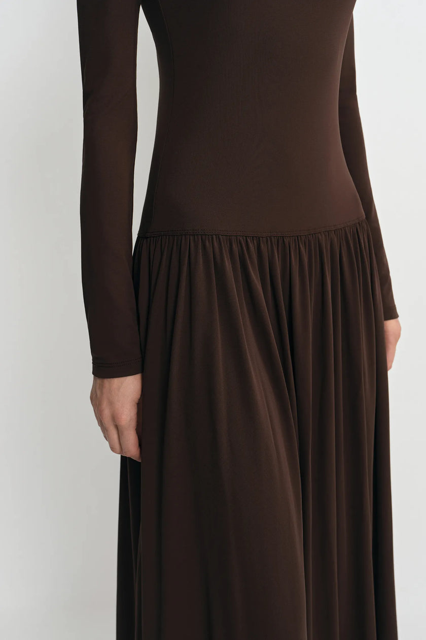 A close-up of a model wearing a dark brown long-sleeve maxi dress with a fitted bodice and a gathered, flowing skirt, shown against a plain light gray background.