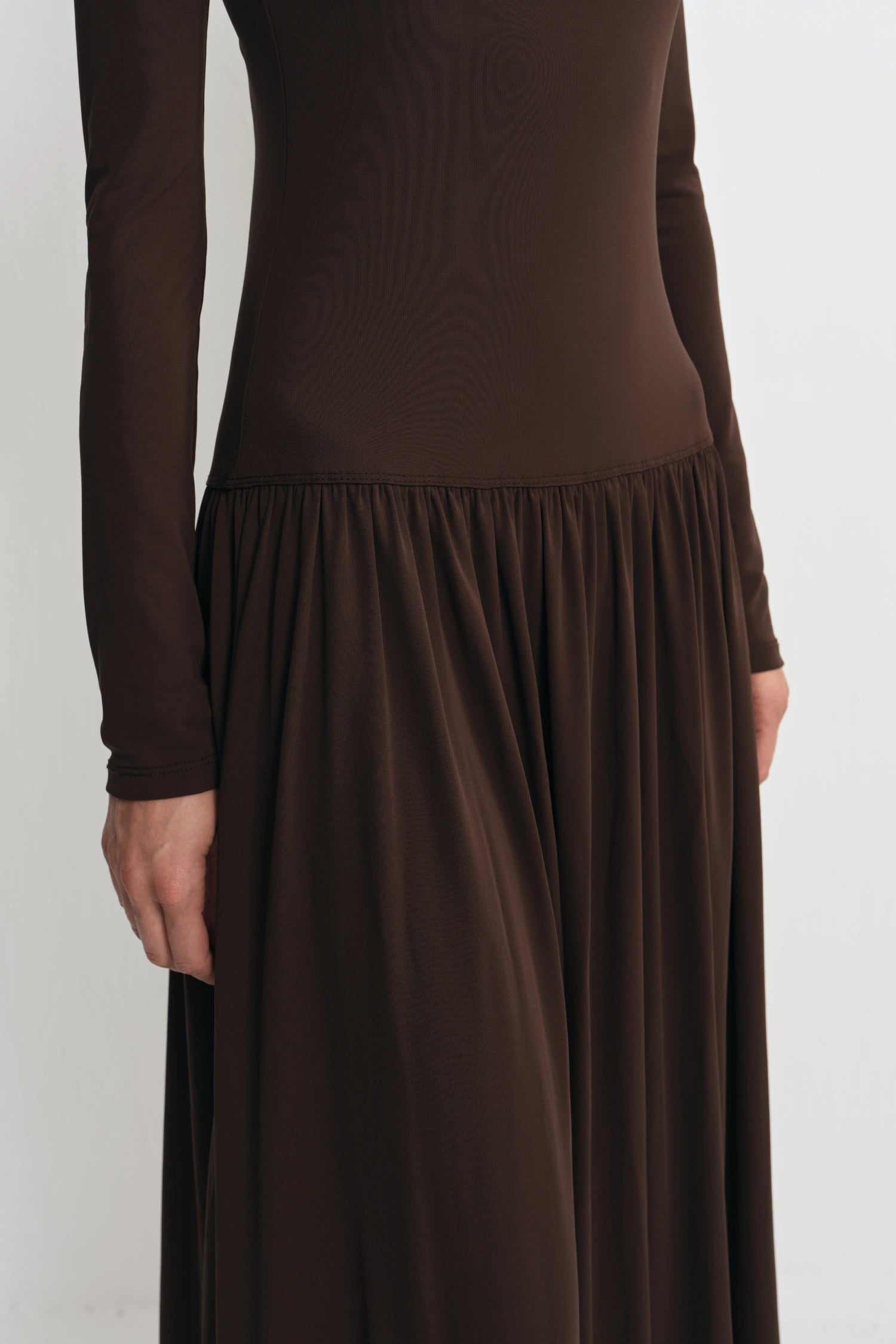 A close-up of a person wearing a long-sleeve brown dress with a fitted bodice and a flowing skirt, shown against a plain light background.