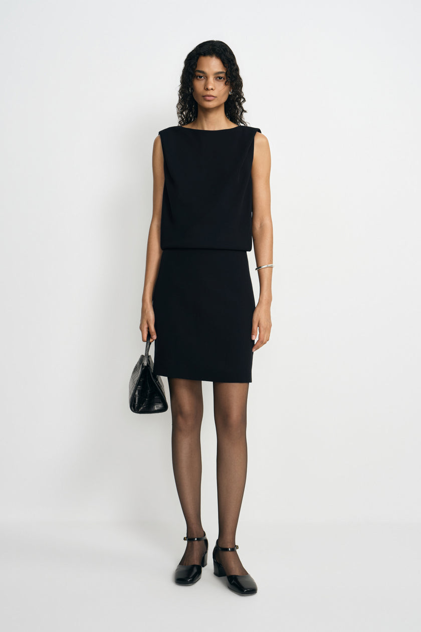 A model facing forward in a black mini dress with a blouson bodice and short straight skirt, carrying a small black handbag and wearing ankle-strap shoes.