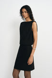 Three-quarter close-up of a model in a black sleeveless mini dress, showing the wide boat neck and folds of the bodice draping over the waist.