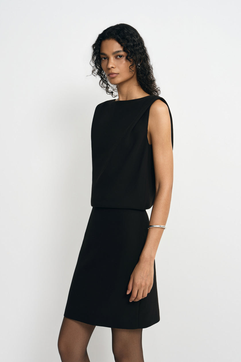 Three-quarter close-up of a model in a black sleeveless mini dress, showing the wide boat neck and folds of the bodice draping over the waist.