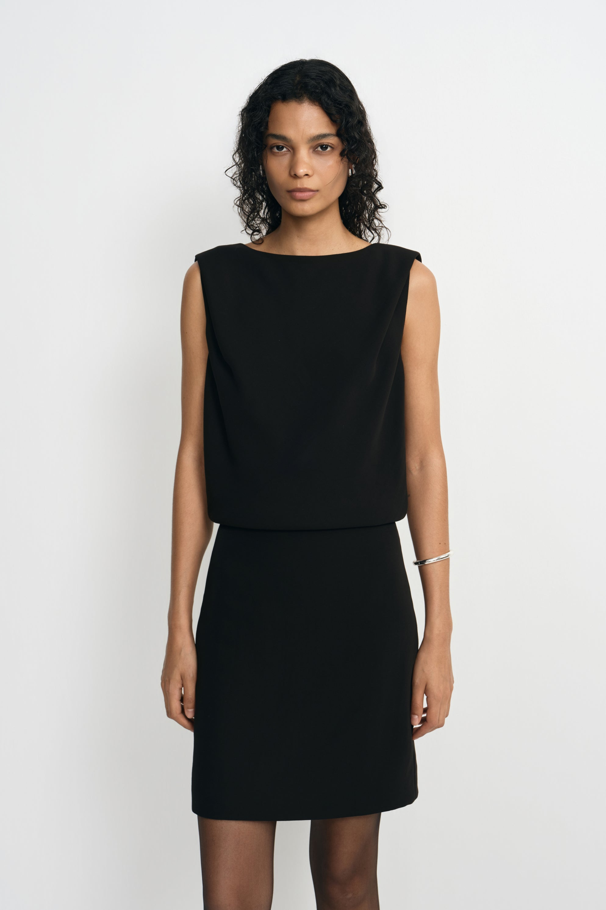 Front detail of a black mini dress on a model, showing the boat neckline, loose bodice overlapping the waist and straight short skirt.