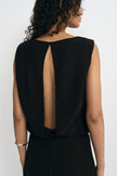 Close-up of a black sleeveless dress on a model from behind, showing the long open-back cutout framed by draped panels and fastened at the neckline.