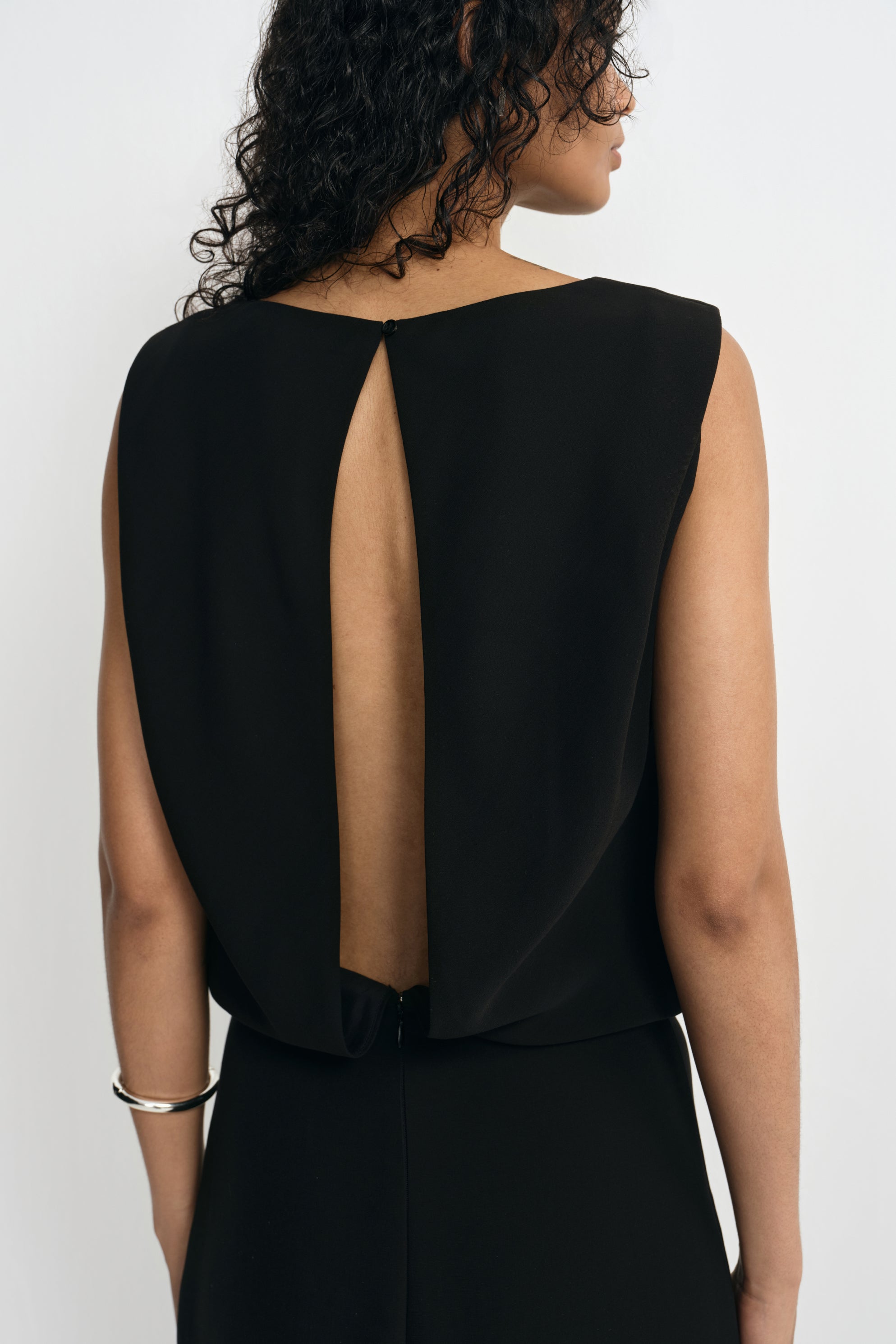 Close-up of a black sleeveless dress on a model from behind, showing the long open-back cutout framed by draped panels and fastened at the neckline.