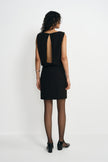 Back view of a model in a black mini dress with an opening from neckline to waist, paired with black ankle-strap shoes below the short skirt.