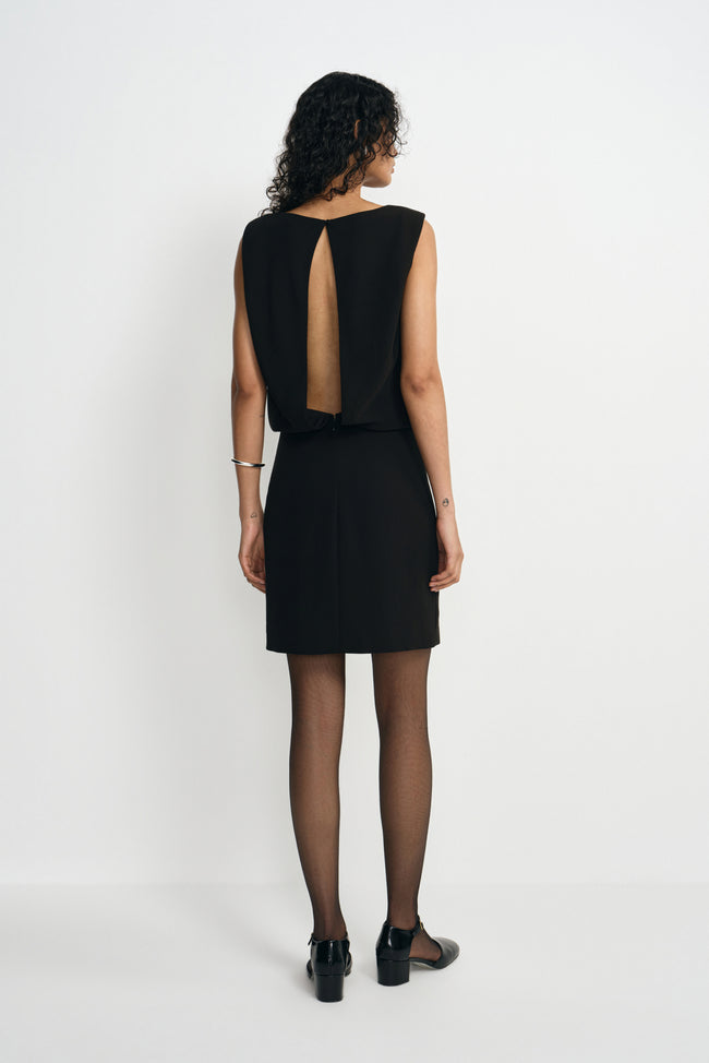 Back view of a model in a black mini dress with an opening from neckline to waist, paired with black ankle-strap shoes below the short skirt.