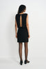 Back view of a model in a black mini dress with an opening from neckline to waist, paired with black ankle-strap shoes below the short skirt.