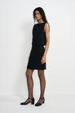 A model standing at an angle in a black sleeveless mini dress with a draped bodice and straight skirt, paired with black block-heel ankle-strap shoes.